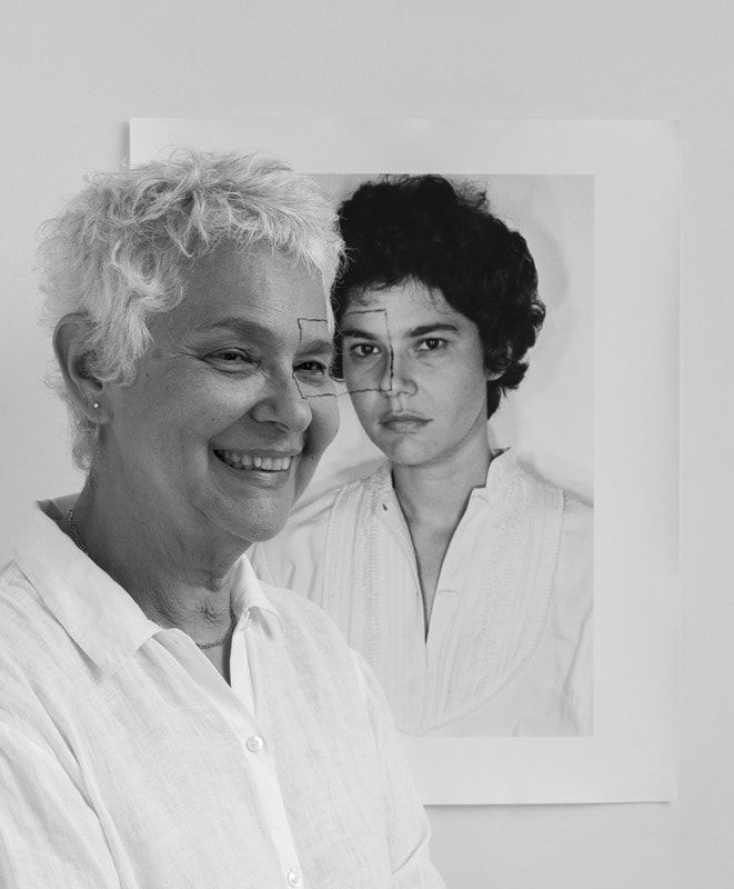 A greyscale photograph of a Latin American, white-haired, older woman standing and smiling (left) with a box drawn around her left eye. A photo of a young Latin American woman with short, dark curly hair staring defiantly at the camera hangs on the wall (right). The young woman has a box drawn over her right eye, which connects to the box around the older woman’s left eye.
