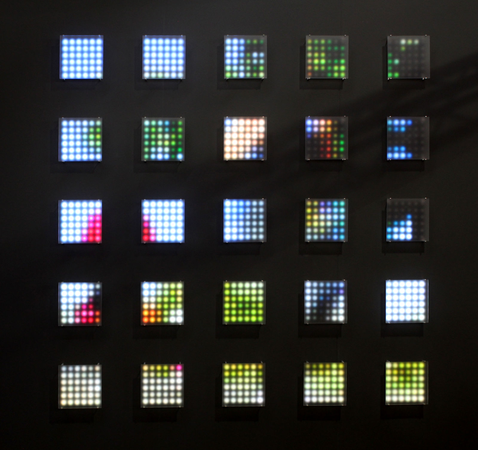 Still image of a spread out grid of twenty five square pieces with multi-colored LEDs behind Plexiglass. The LEDs display pixelated scenes of runners from a marathon