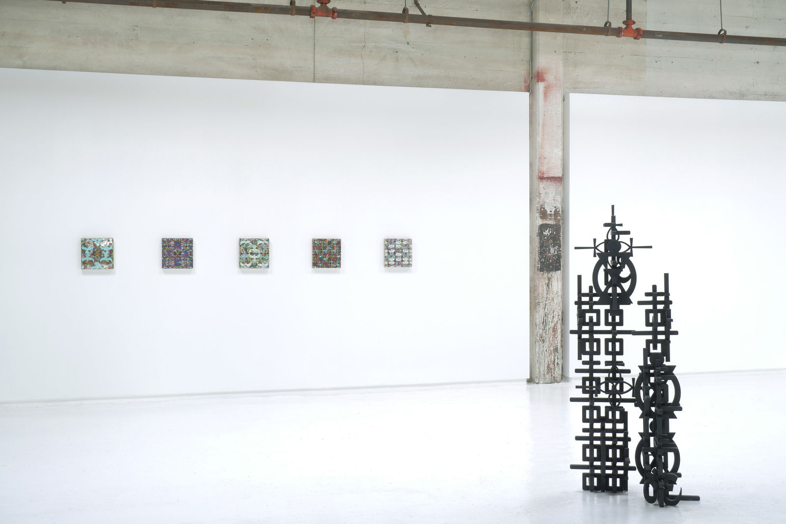 Installation view of Totem with 5 square panel paintings hanging in a horizontal line on the wall behind the sculpture
