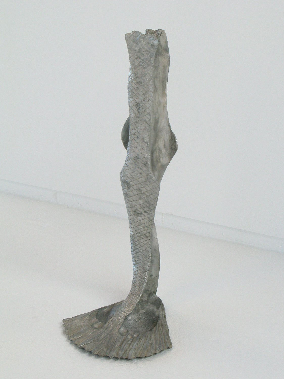 Aluminum mold of the negative space between the artist's legs which has been textured to resemble fish scales and a tail fin. At the base of the sculpture where the tail fin takes shape imprints of the artist’s feet are visible