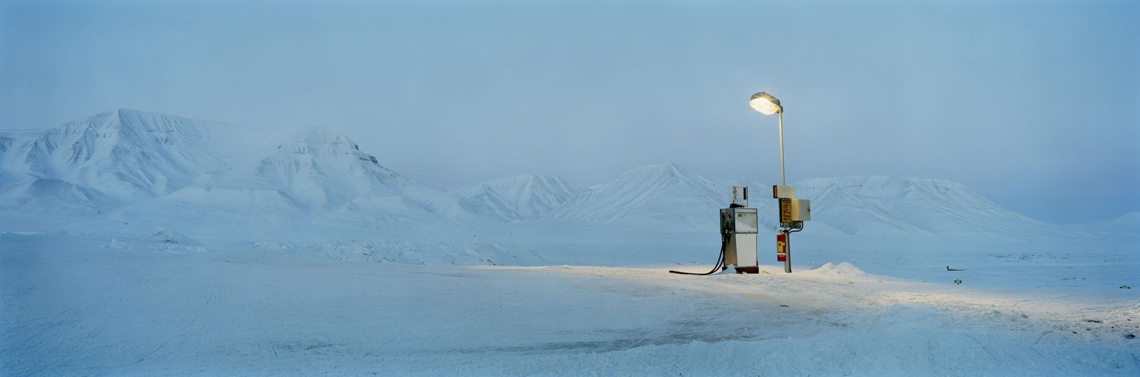 Digital panoramic landscape photograph of a singular petrol pump underneath a shining overhead light The illuminated pump contrasts with the mountainous blue arctic landscape behind it