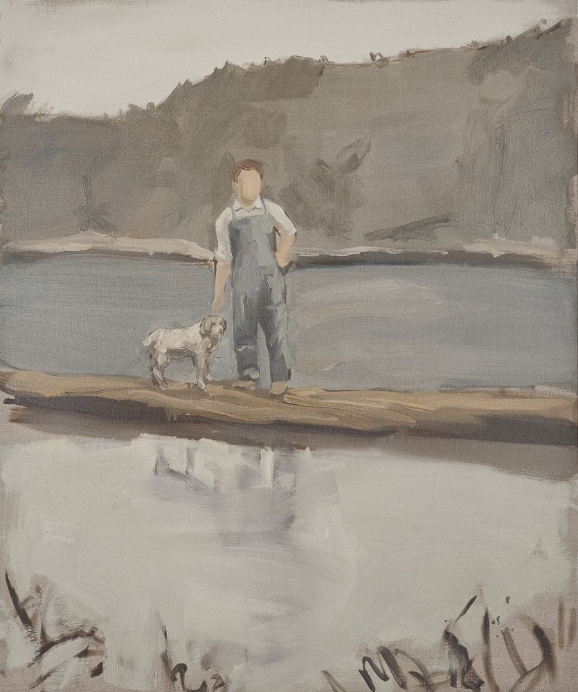 Faceless man in overalls with dog stands on dock in lake with gray mountains in the background.