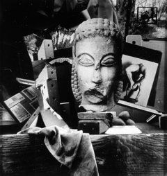 Black and white collage of items including sculpture of a figure’s head, a nude polaroid, a piece of cloth and children’s toys all resting on a wood surface.