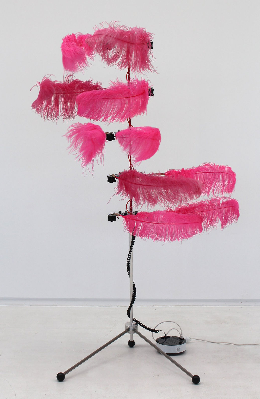 Tall silver pole with five large neon pink feathers on either side that move in all sorts of patterns. There are red and black cords wrapped around pole leading to two black poles at the base spread out for support.
