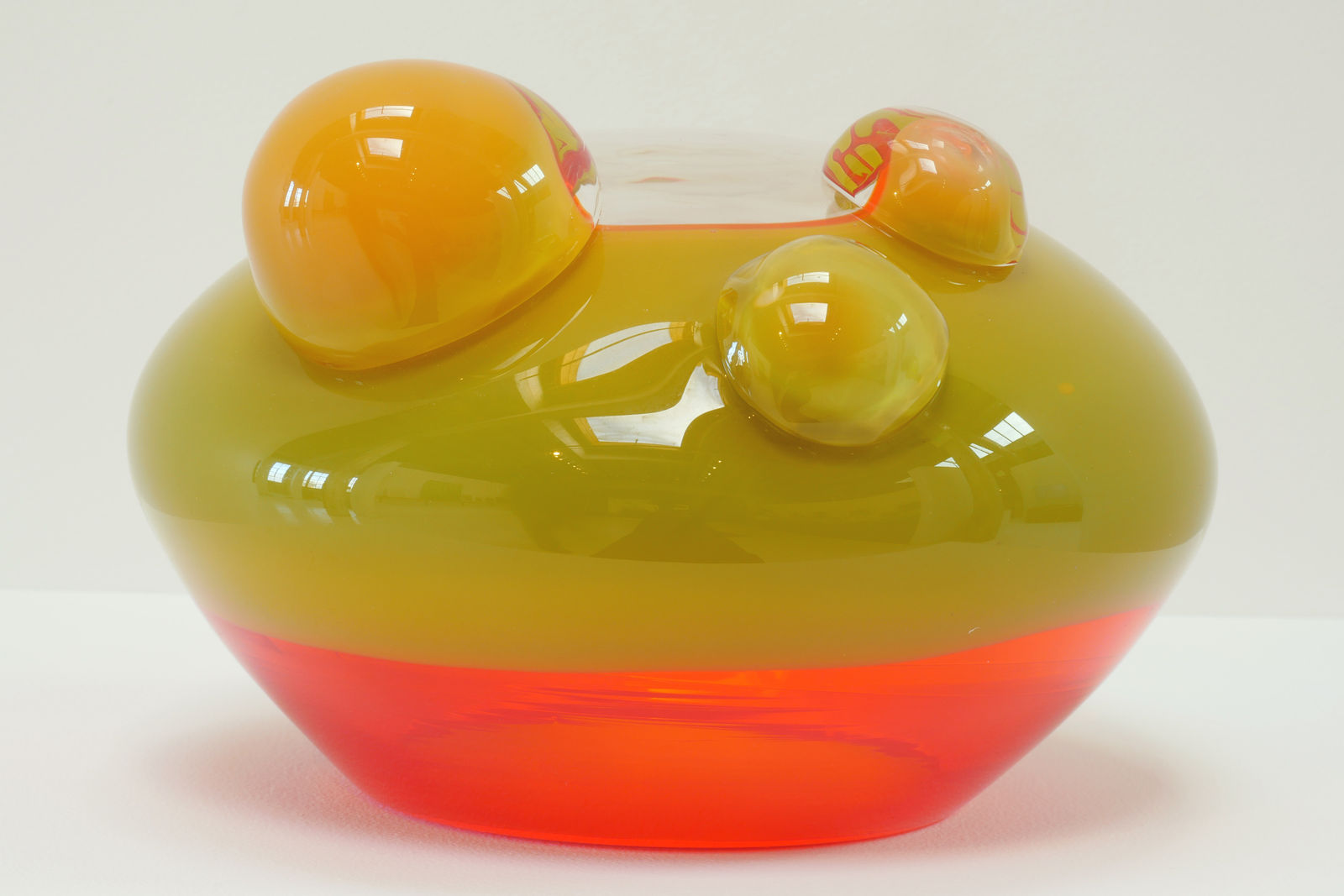 A UFO-like glass sculpture. Top 2/3 olive colored, bottom 1/3 fluorescent orange. Three bubbles of differing sizes adorn the top.