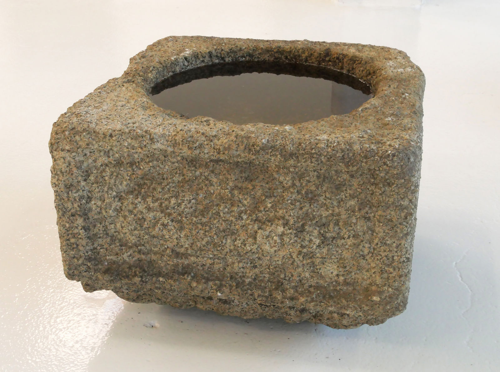 Side detail of weathered square stone basin with circle in center holding water.