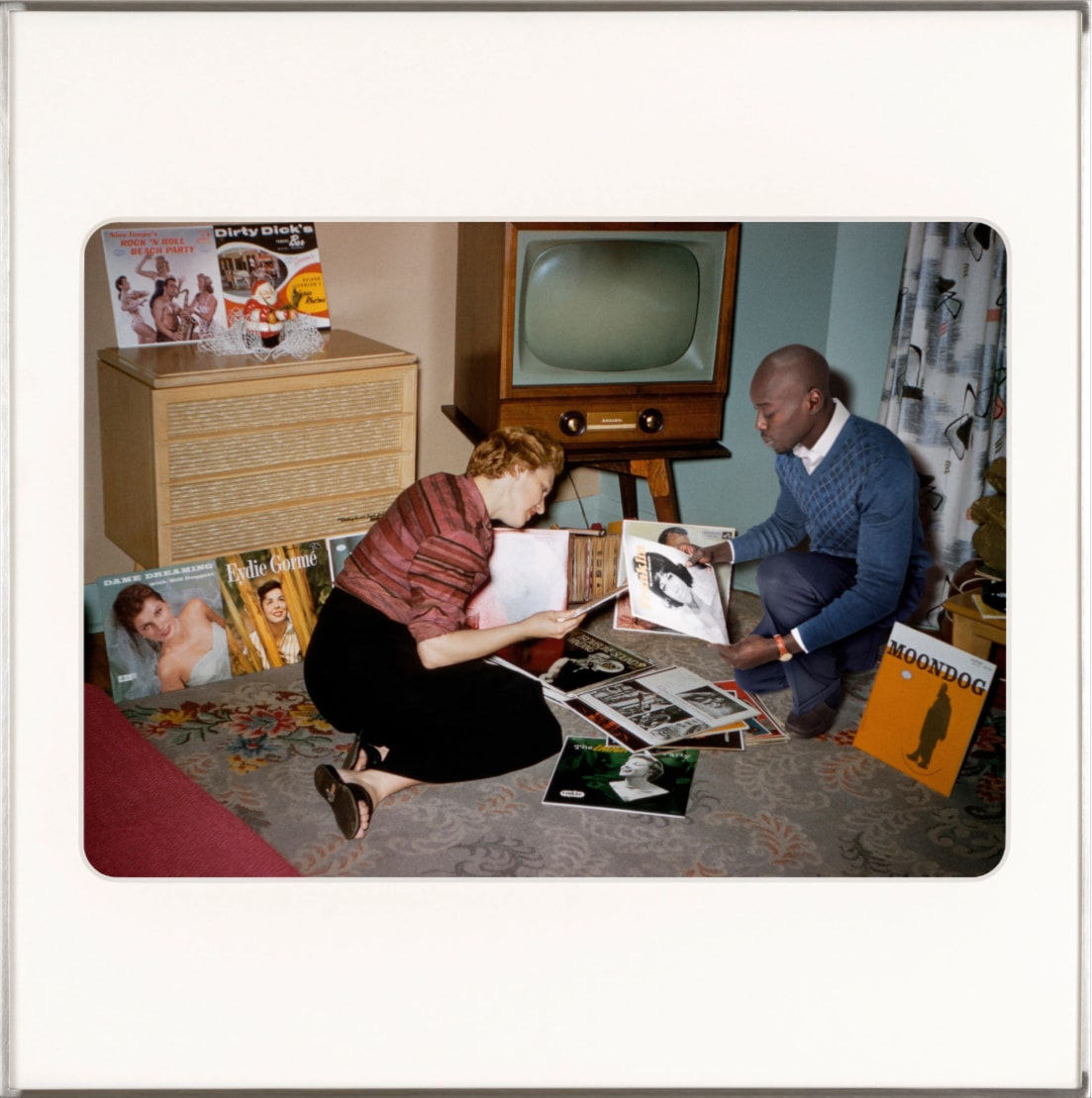 Artist Omar Diop sitting on a living room floor looking at vinyl with a white woman. By Lee Shulman & The Anonymous Project.
