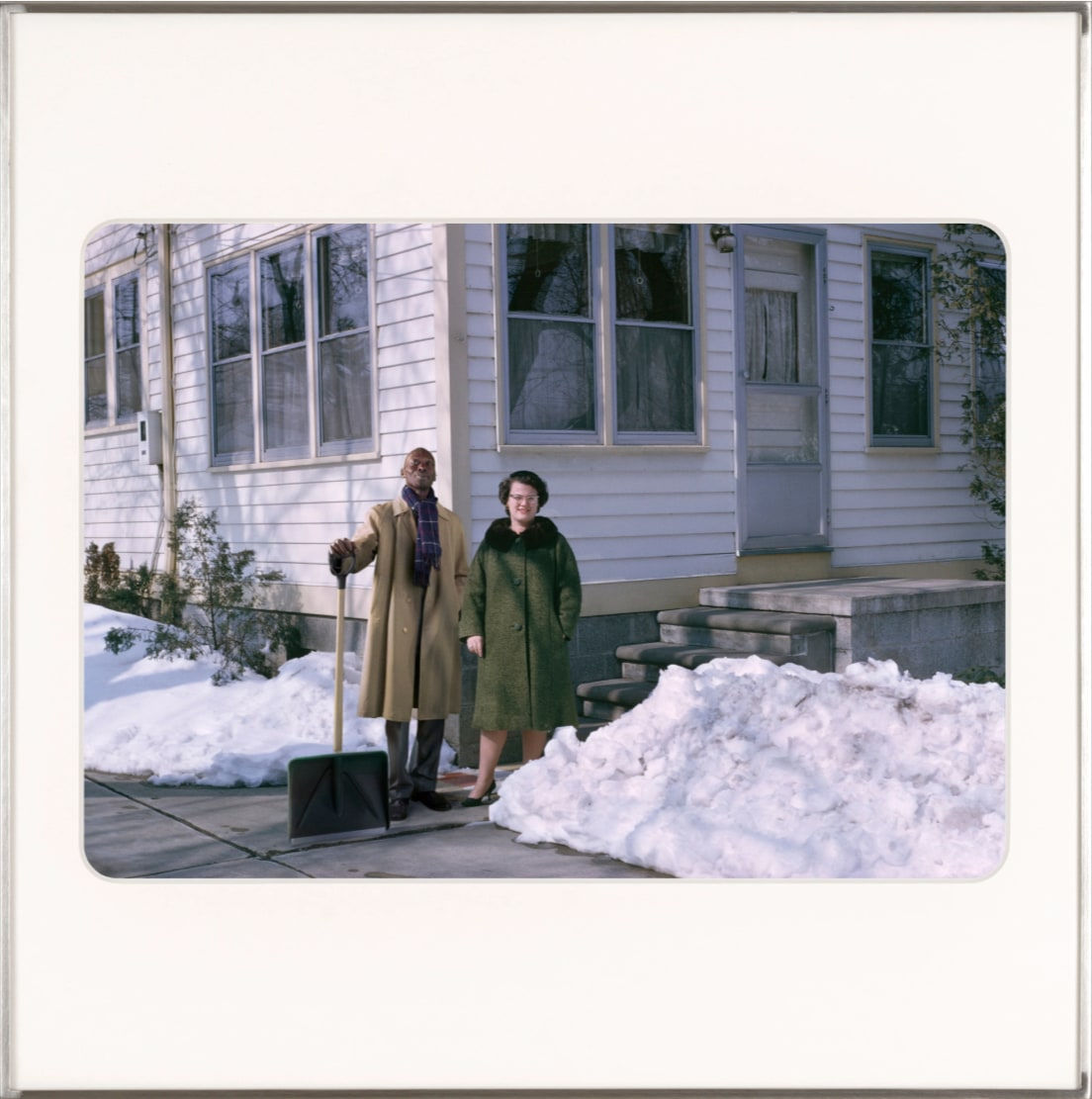Artist Omar Diop standing in a coat with a shovel next to a white woman. Both are in front of a white paneled house in the snow. By Lee Shulman & The Anonymous Project.