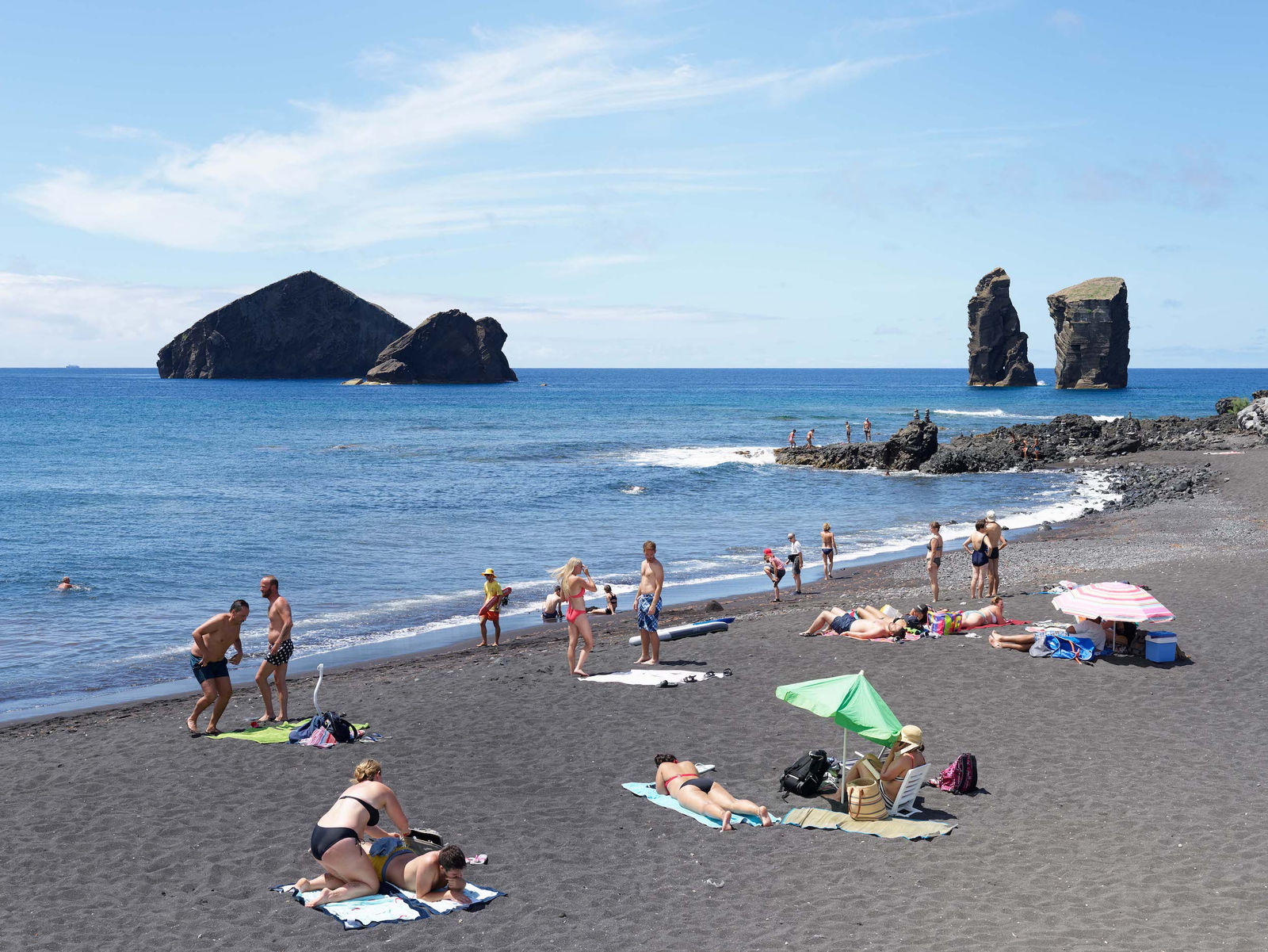 Photograph by Massimo Vitali showing beachgoers relaxing on a black sand beach with rocky sea stacks rising from the blue ocean in the background.