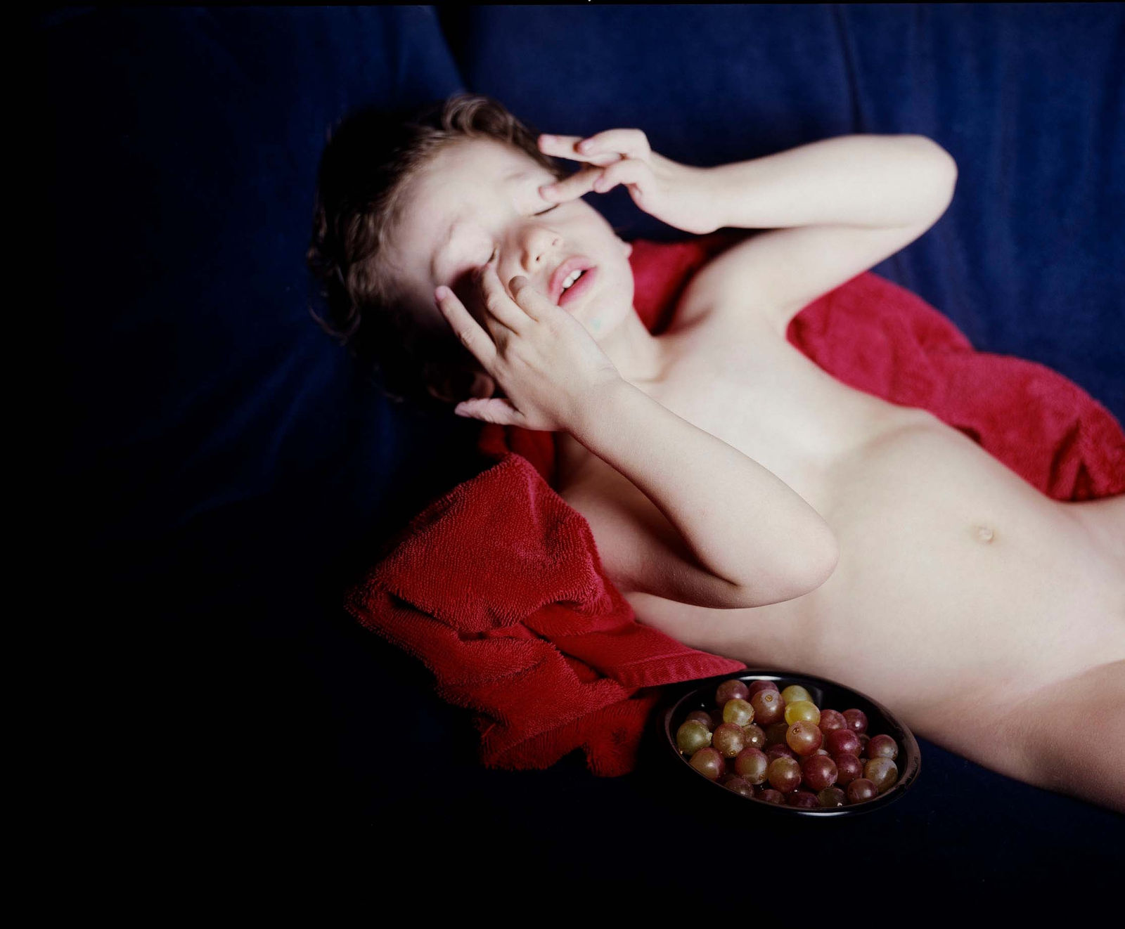 Elinor Carucci photograph of her child laying on a red blanket with a bowl of grapes and covering their eyes