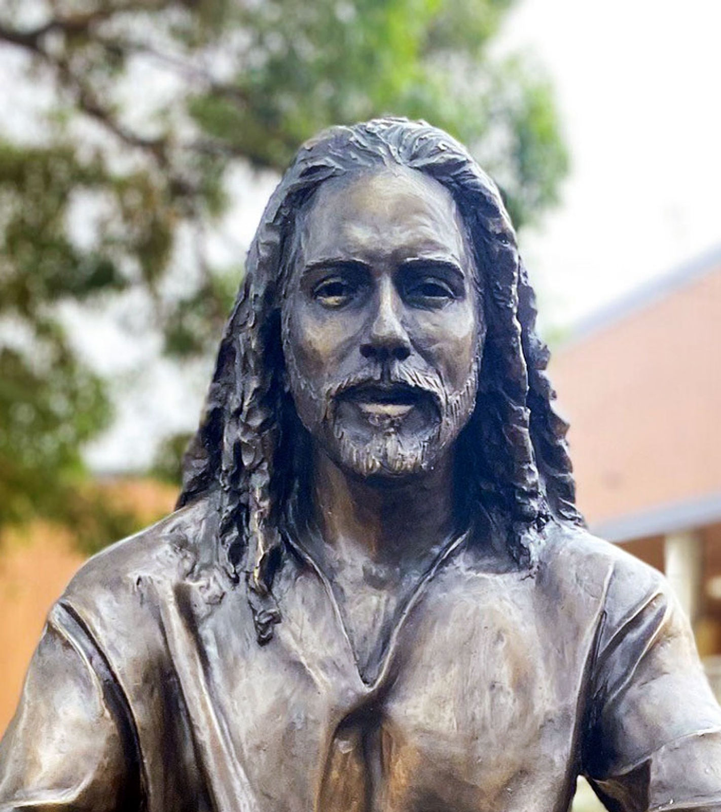 A close-up of the bronze statue depicting Jesus with detailed facial features, including flowing hair and a beard, set against a natural background with trees in soft focus.