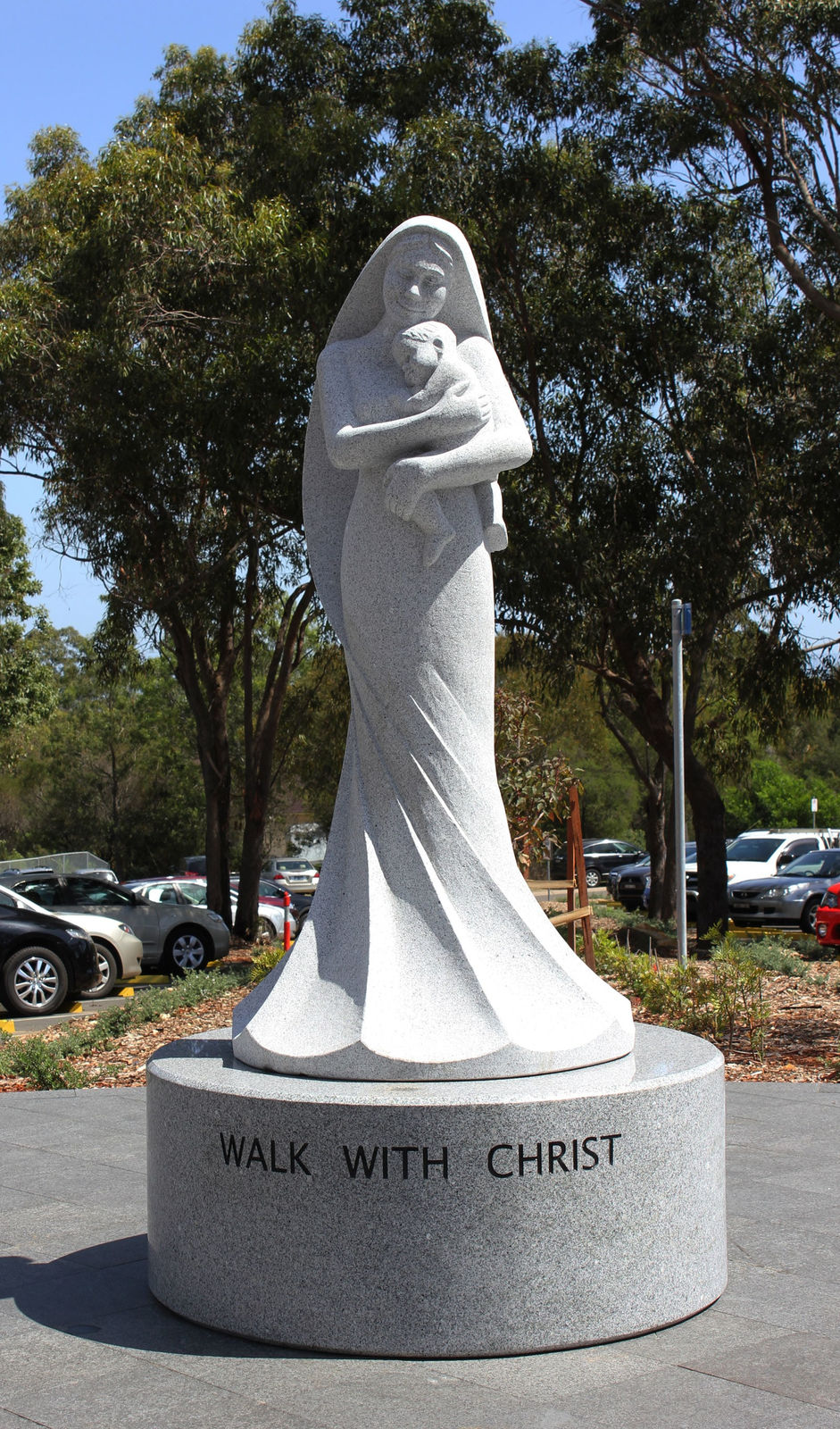 A smooth granite statue of a serene woman holding an infant close to her chest, standing on a circular base with the words 