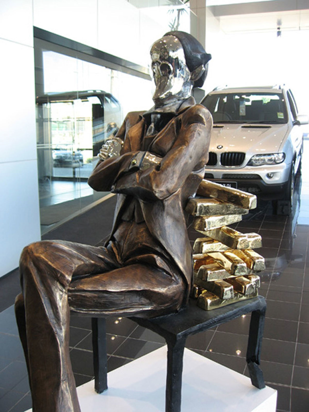 The large banker sculpture showcased in a car dealership, with luxury vehicles in the background. The figure, seated on a chair with gold bullion forming the backrest, conveys themes of wealth and affluence. The polished reflective face adds a modern, surreal touch to the piece.