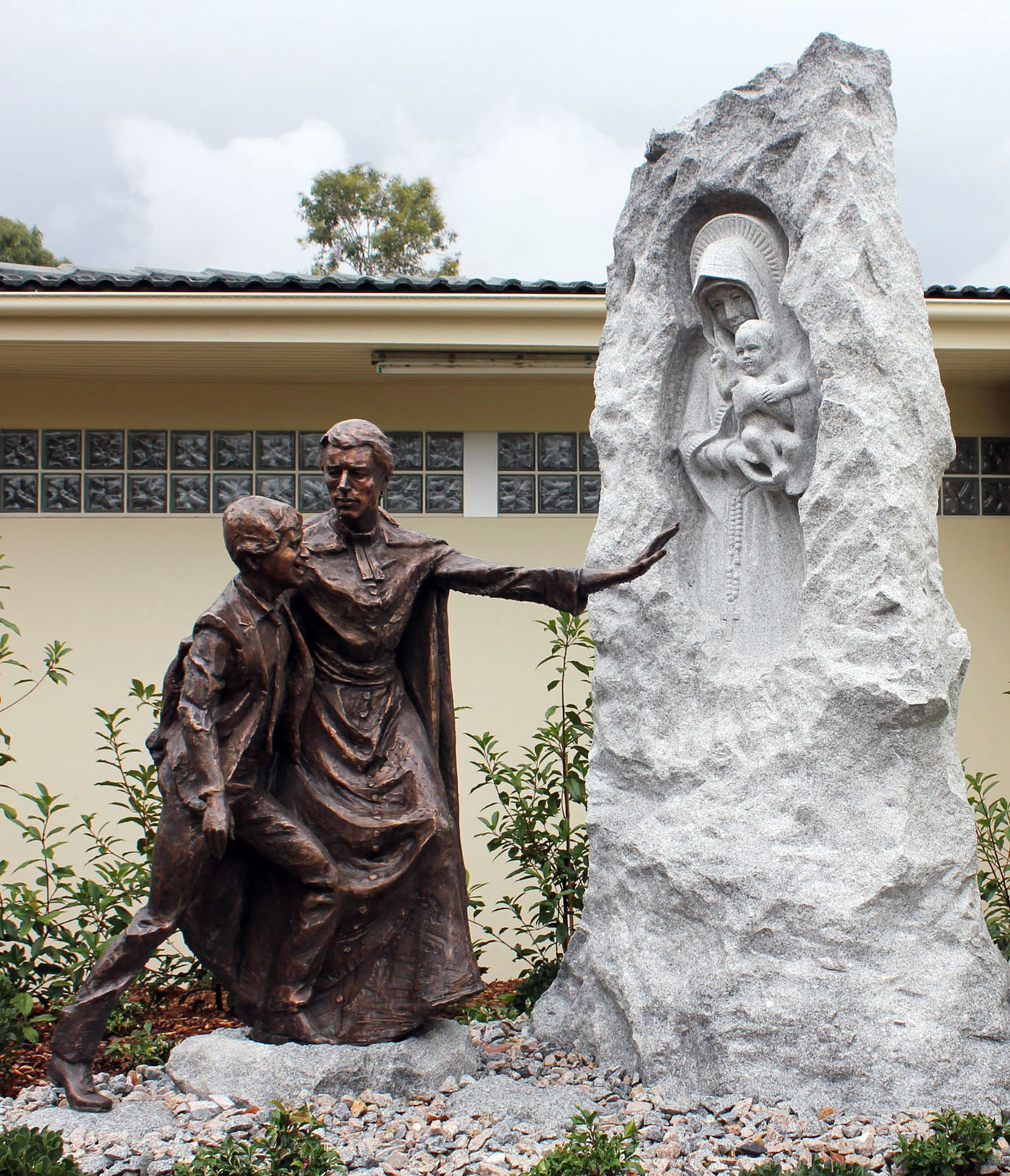 Bronze statue of St. Marcellin Champagnat guiding a young student through the mountains, pointing toward a granite relief depicting an apparition of the Virgin Mary holding the Christ child. The composition symbolizes faith, perseverance, and divine inspiration during their journey, blending the textures of bronze and granite to enhance the spiritual narrative.