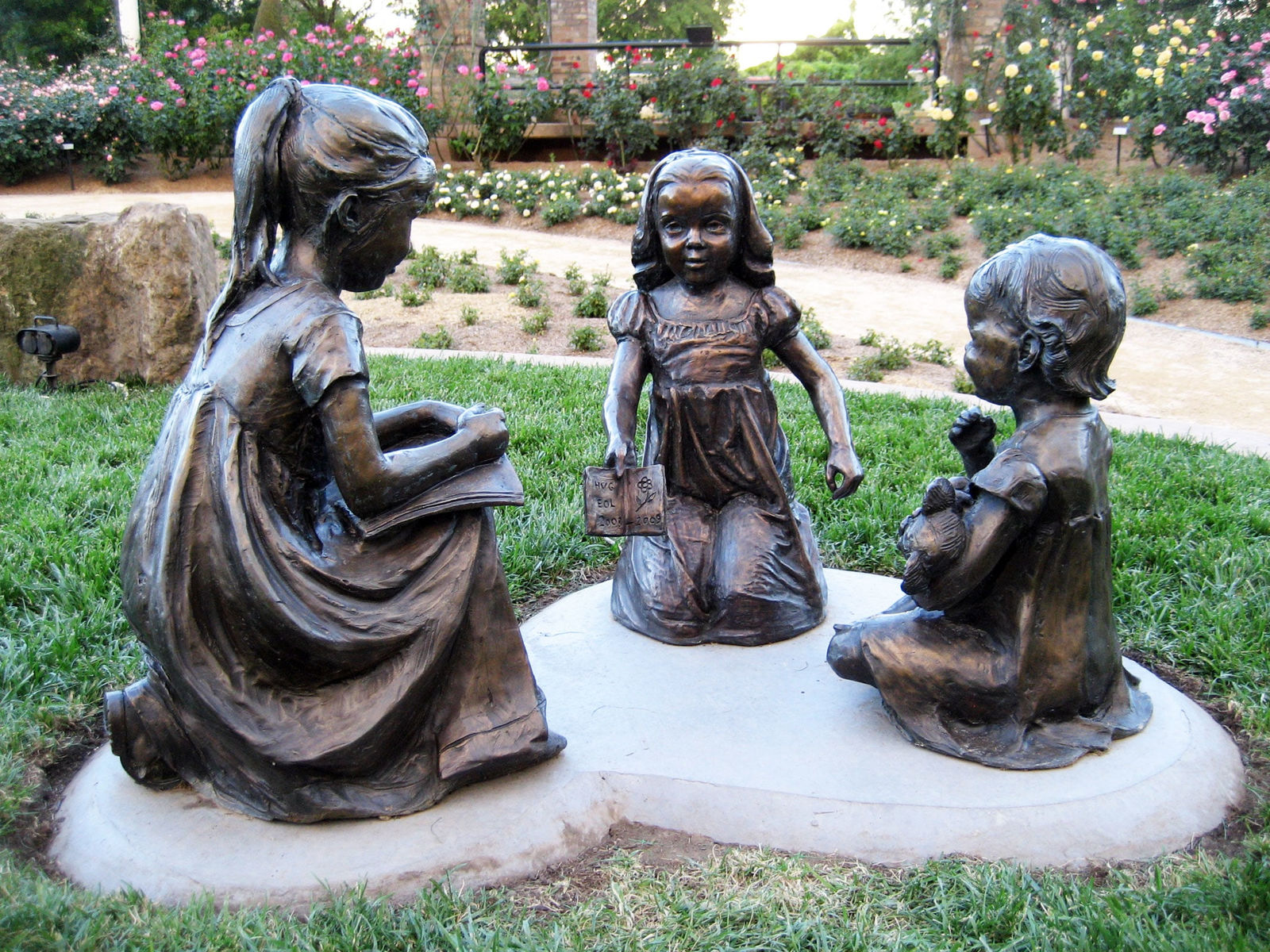 Bronze sculptures of three young girls seated in a circle on a grass-covered garden pathway. One girl holds an open book, another clutches a teddy bear, and the third engages attentively, creating a serene and playful storytelling scene. The setting features vibrant rose bushes and a tranquil garden backdrop.