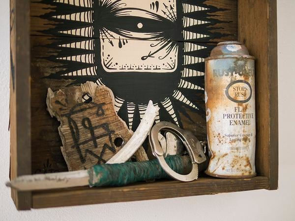 Wooden shelf with painting of an eye by GATS with several trinkets in the shelf including a spray can, handcuffs, piece of wood and a knife