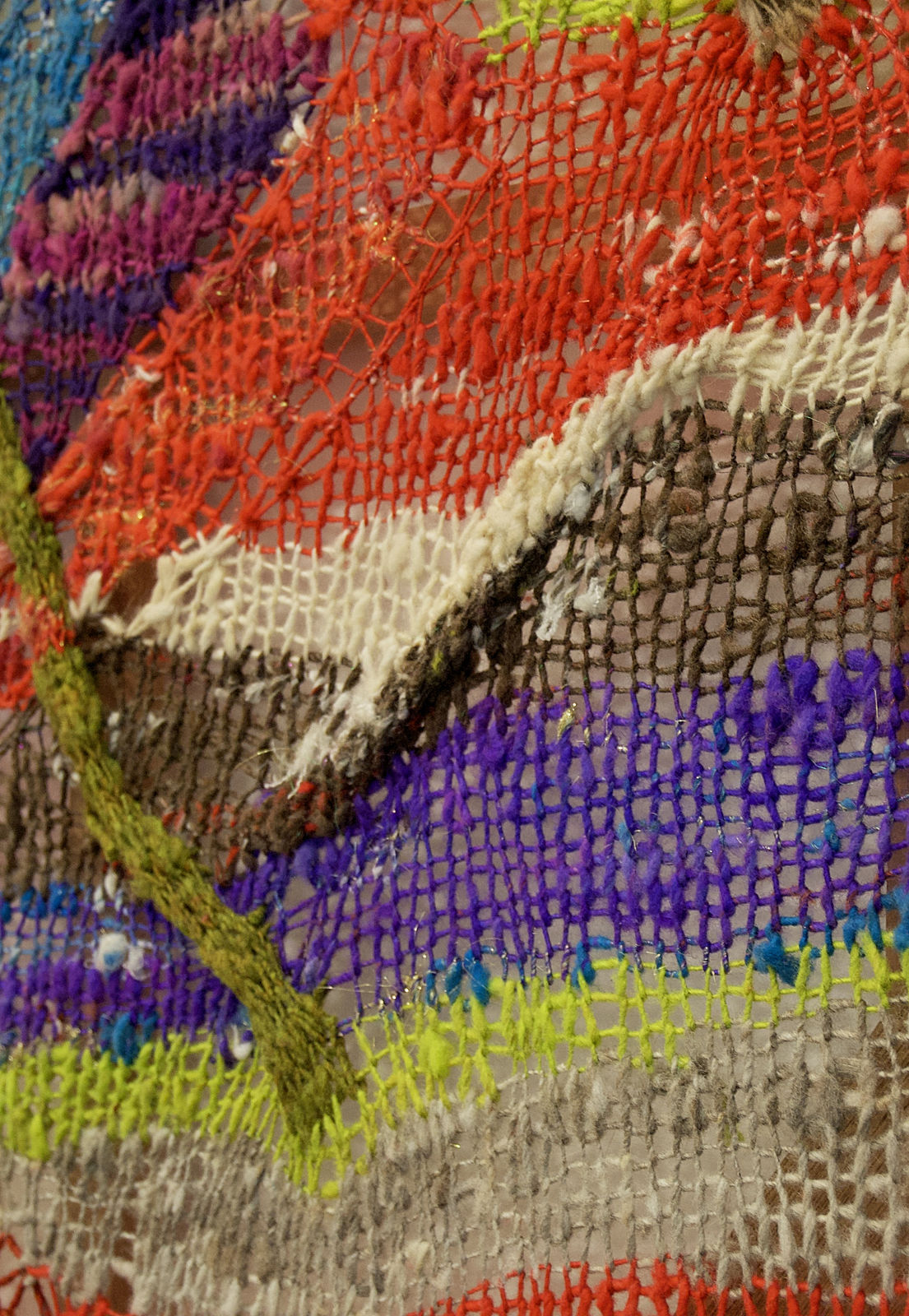 A close-up detail of a striped, diagonal composition made from multi-colored fibers stretched across a redwood frame as one would stretch a painting. The fibers are knit at varying thicknesses so that in places one can see through the stitches to the wall behind. The pattern is geometric with two axe-shaped designs rising up from the rest of the picture