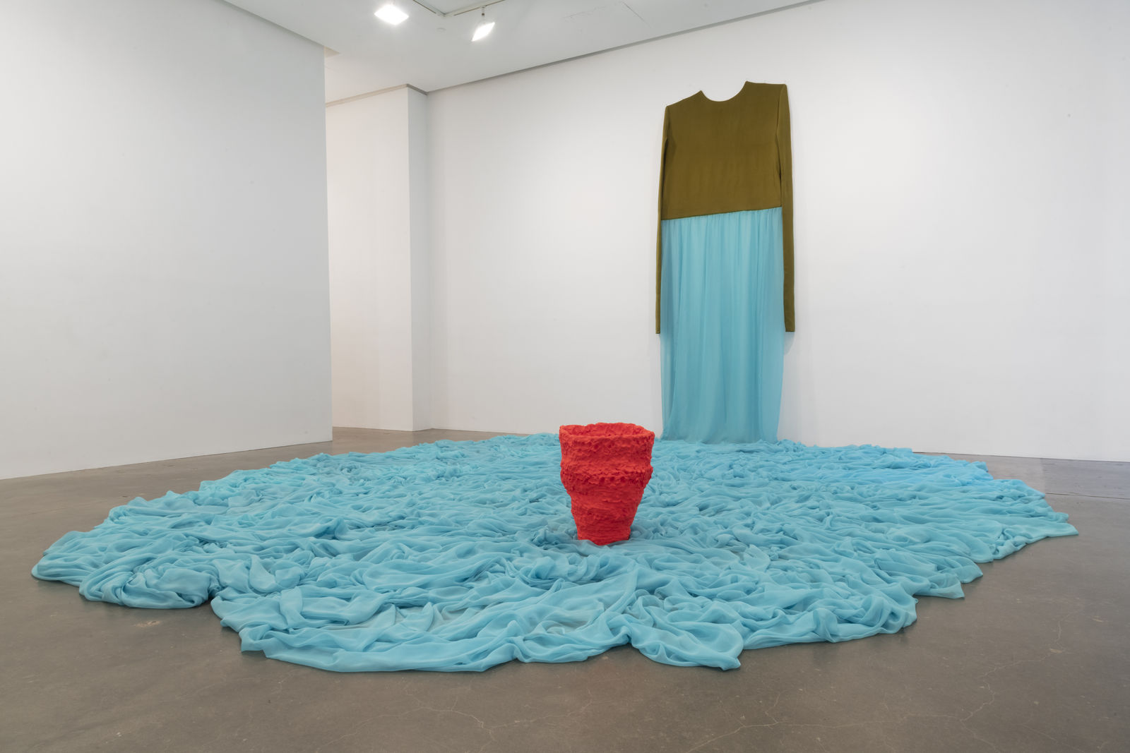 A giant dress made from teal fabric hung on the wall cascading to the floor with a red pot