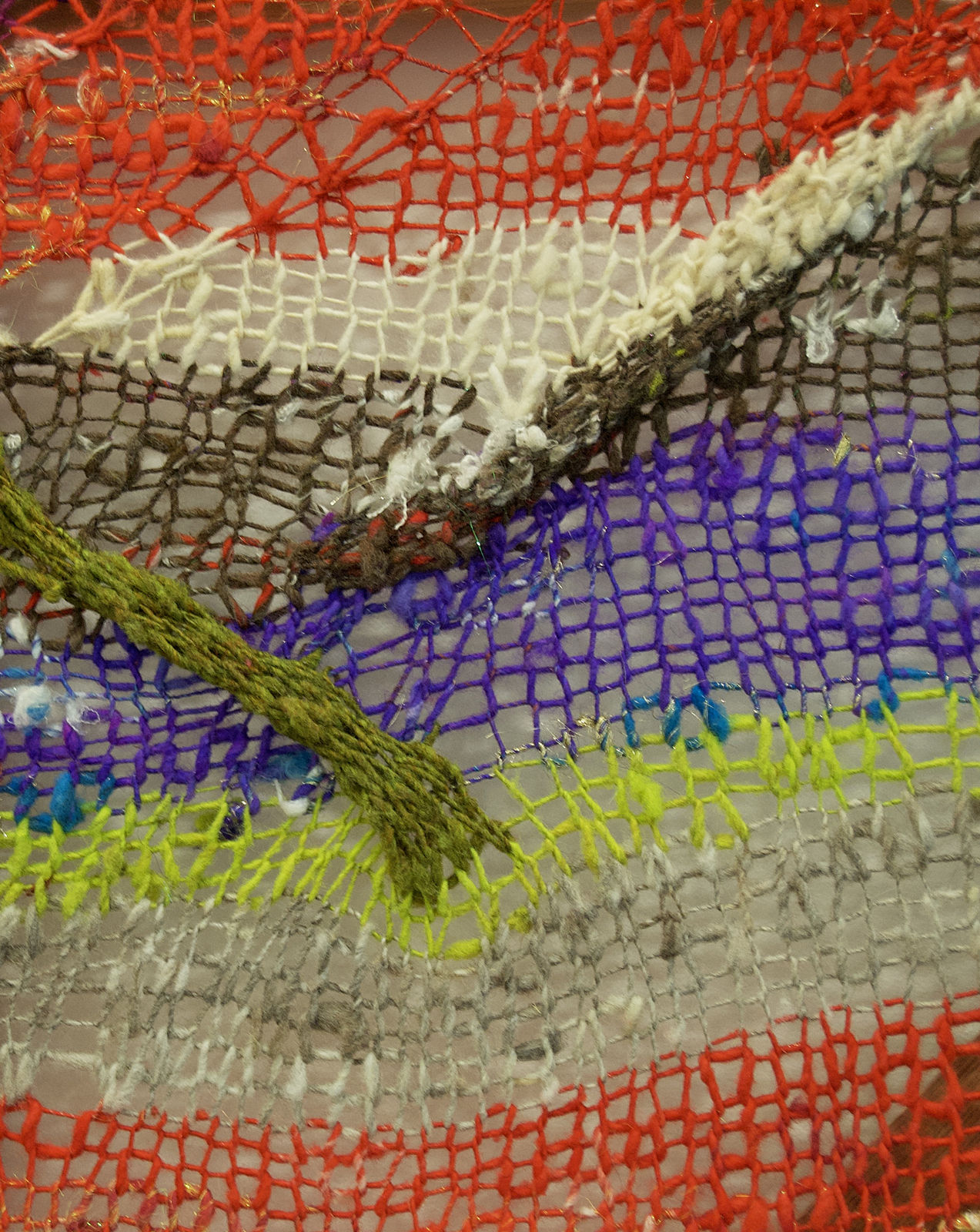 A close-up detail of a striped, diagonal composition made from multi-colored fibers stretched across a redwood frame as one would stretch a painting. The fibers are knit at varying thicknesses so that in places one can see through the stitches to the wall behind. The pattern is geometric with two axe-shaped designs rising up from the rest of the picture