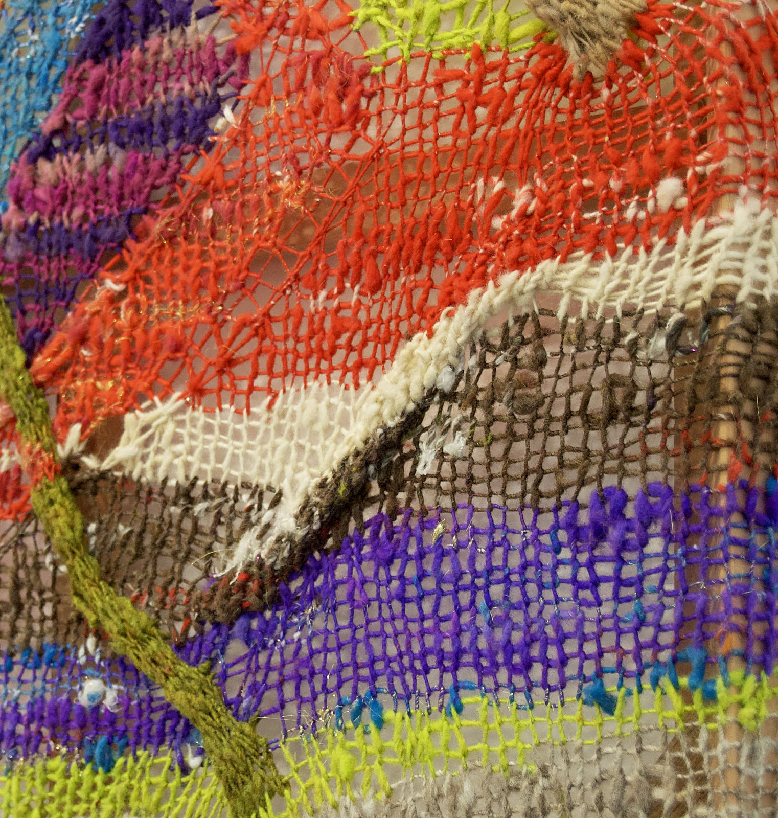 A close-up detail of a striped, diagonal composition made from multi-colored fibers stretched across a redwood frame as one would stretch a painting. The fibers are knit at varying thicknesses so that in places one can see through the stitches to the wall behind. The pattern is geometric with two axe-shaped designs rising up from the rest of the picture