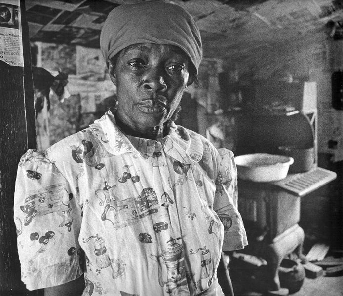Black and white photograph of a woman, Mrs. Pettway, in her kitchen with walls covered in newspapers featuring Martin Luther King in Canton Bend, Alabama