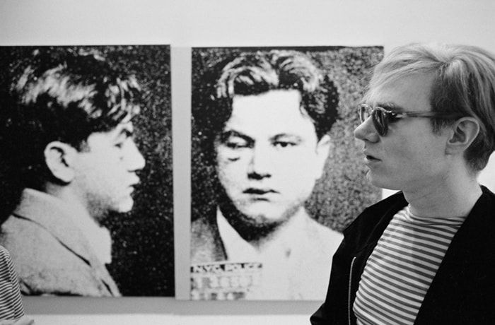 Black and white photograph of Andy Warhol in profile in front of a screen print of a man’s mug shot