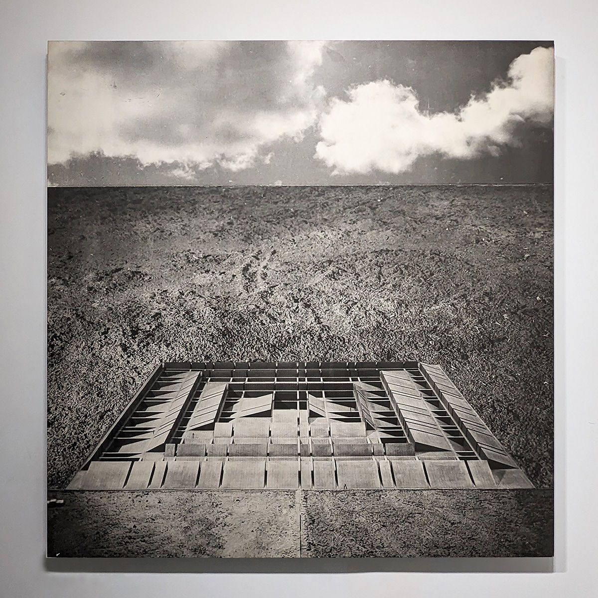 Black and white photomontage of a flat landscape with a three dimensional structure in the form of raised grid lines, viewed from above and at a distance