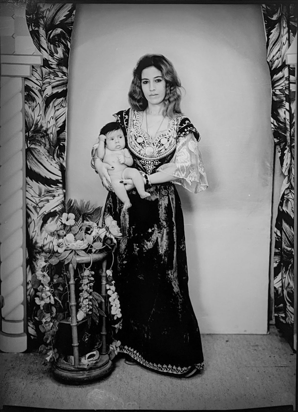 1960s black and white photograph of a woman standing wearing an embroidered dress holding a baby in her arms