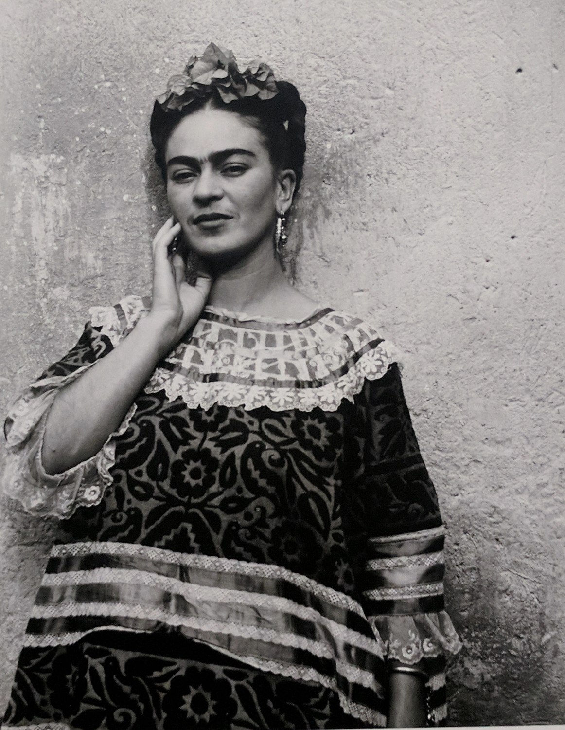 Black and white photograph of Frida Kahlo in traditional Tehuana dress leaning against the exterior wall of the Blue House in Mexico City with her hand grazing her face
