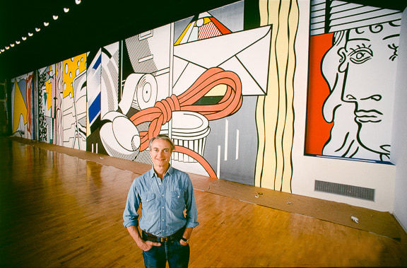 Photograph of Roy Lichtenstein smiling in front of a large mural featuring a woman’s face and an array of household objects
