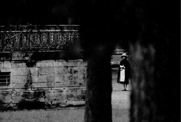 Black and white photograph of Designer Coco Chanel walking in a garden as seen through a small opening of a gate