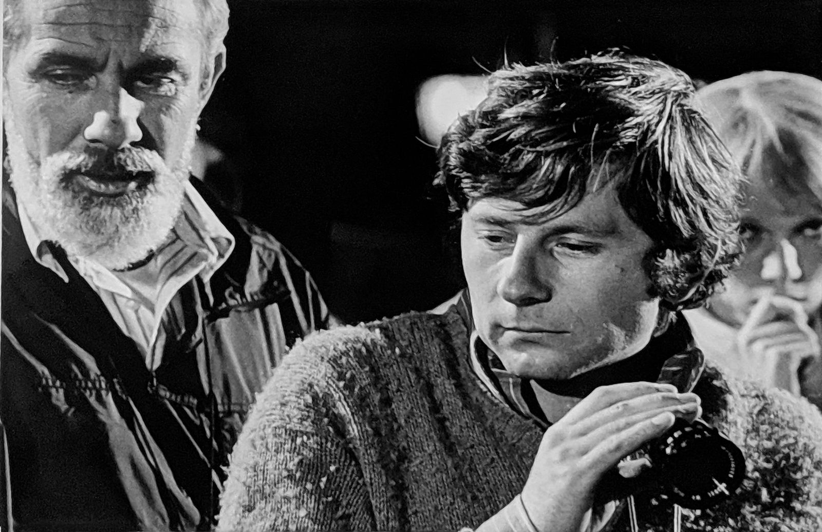 Black and white photograph of William A. Fraker, Roman Polanski and Mia Farrow looking at them in the background, New York City, on the set of “Rosemary's Baby”