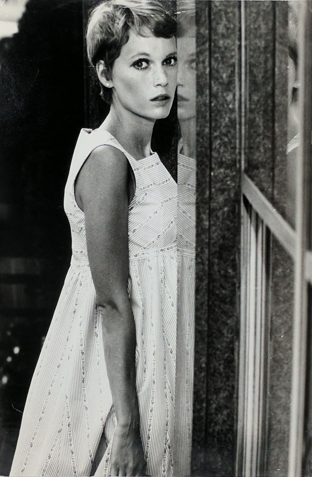 Black and white photograph of Mia Farrow on the set of “Rosemary's Baby”, wearing a white dress and looking directly at the camera