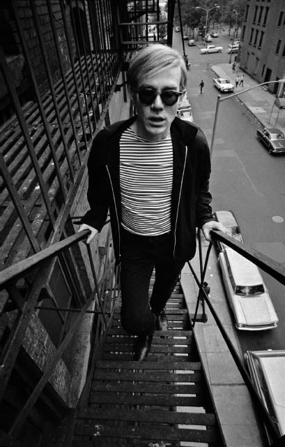 Black and white photograph of Andy Warhol in a Breton striped shirt and sunglasses ascending a fire escape above a city street