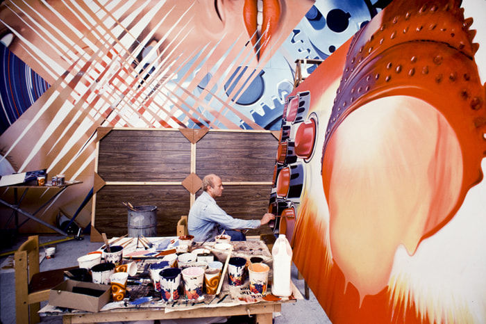 Photograph of James Rosenquist sitting in an artist studio working on a large multicolored painting behind a table with pots of paint