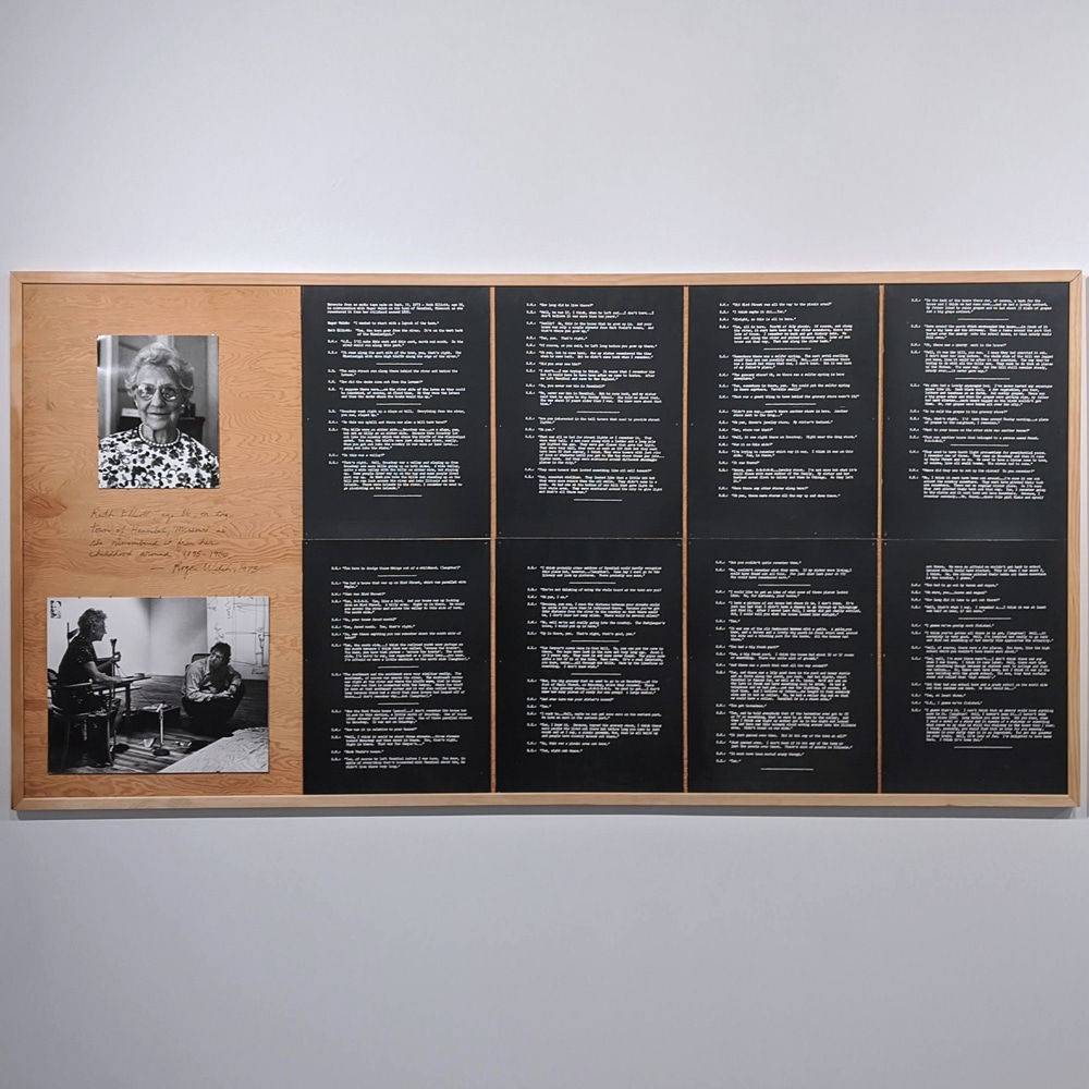 Two photographs of an elderly woman, mounted on wood next to eight pages of white text on black paper arranged in a 2x4 grid, hanging on a white wall