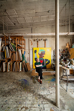 An artist, Carmen Cicero, sits in front of a large figural painting in an industrial loft space filled with stored paintings and materials