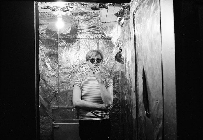 Black and white photograph of Andy Warhol in sunglasses standing in a foil-lined bathroom and looking at the viewer