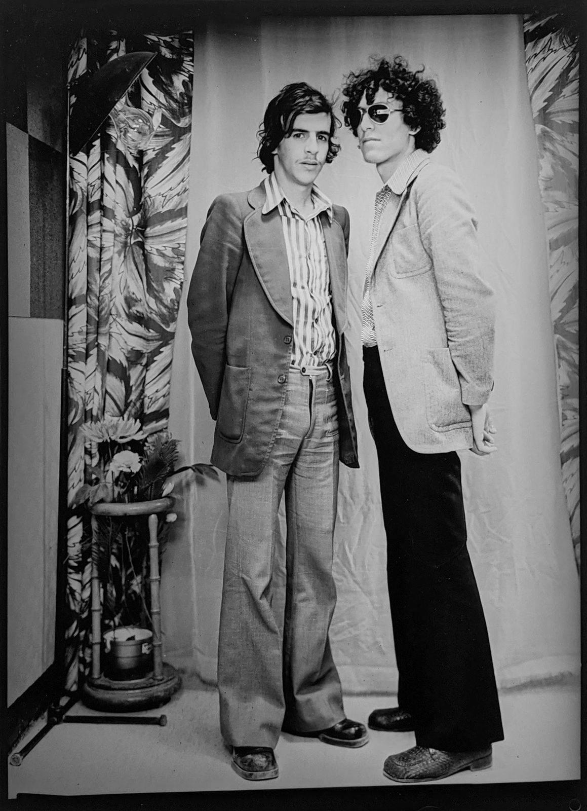 1960s black and white photograph of two men standing, one in profile and one facing the camera, in flared pants and suit jackets