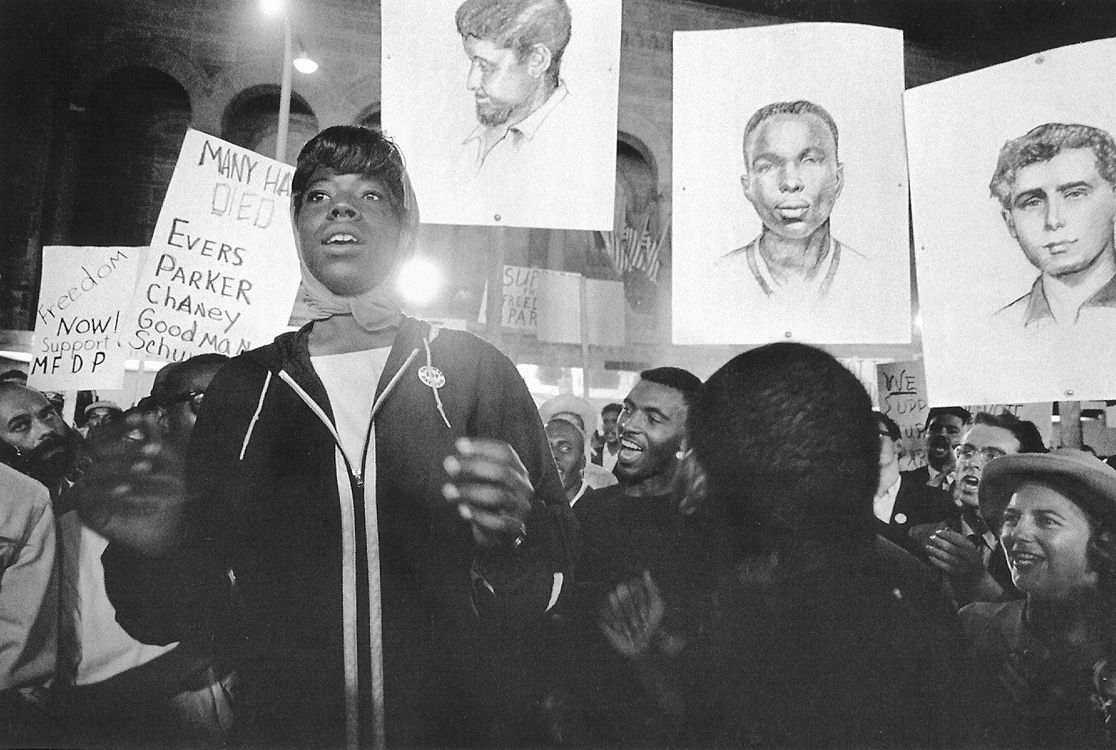 Black and white photograph of a vigil where participants hold posters with images of slain civil rights workers Schwerner, Chaney, and Goodman in Atlantic City, New Jersey