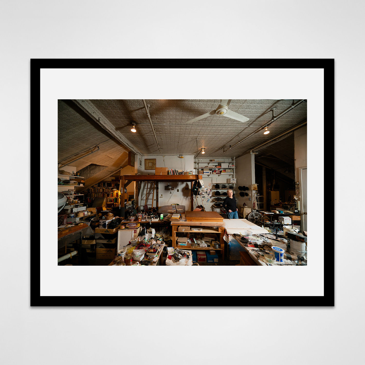 Framed photograph in black wood of an artist, Gilda Pervin, standing amidst an array of work desks, art materials, and books in a loft setting