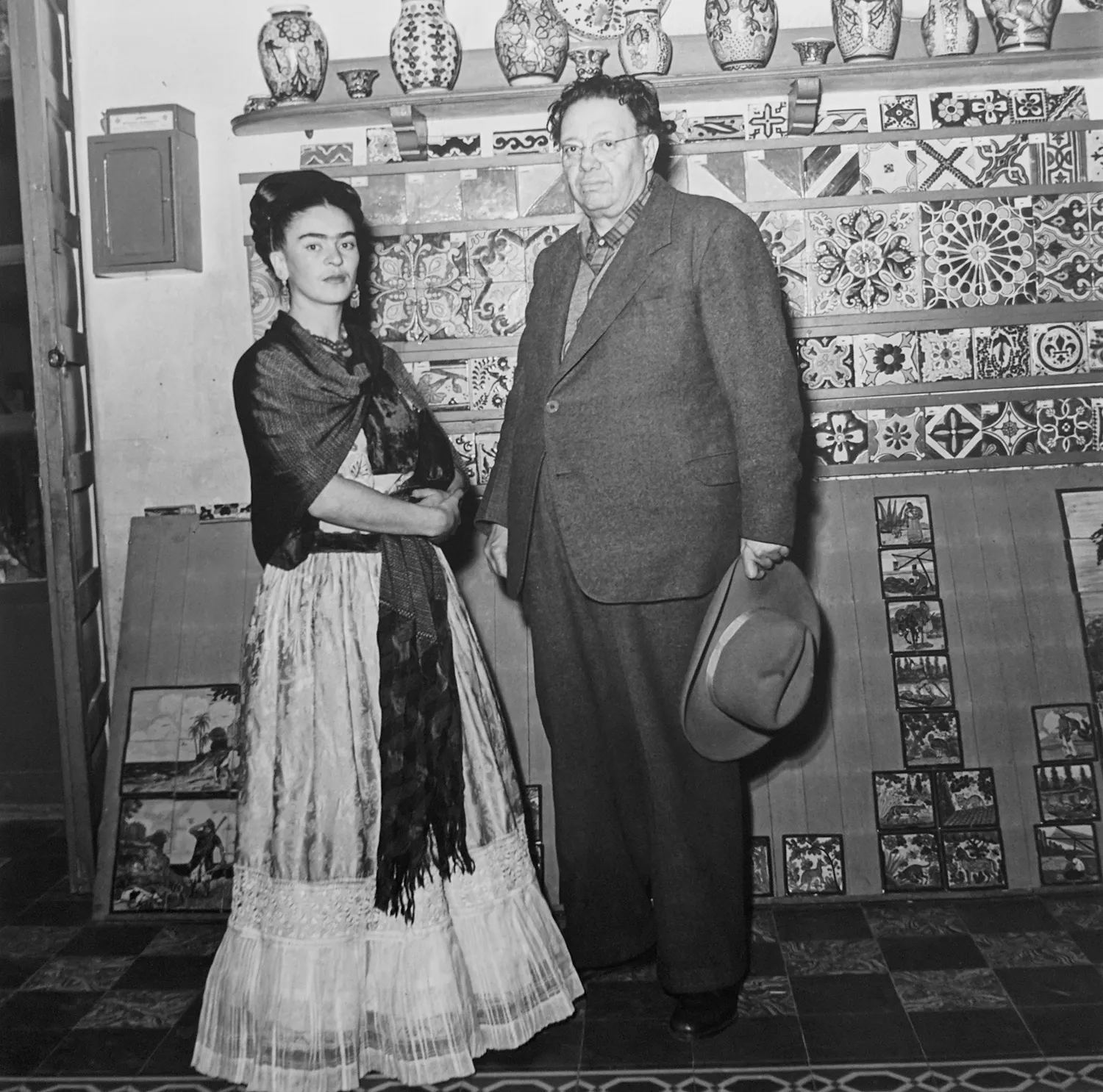 Black and white photograph of Frida Kahlo and Diego Rivera posing in front of a tile-covered wall inside the Blue House