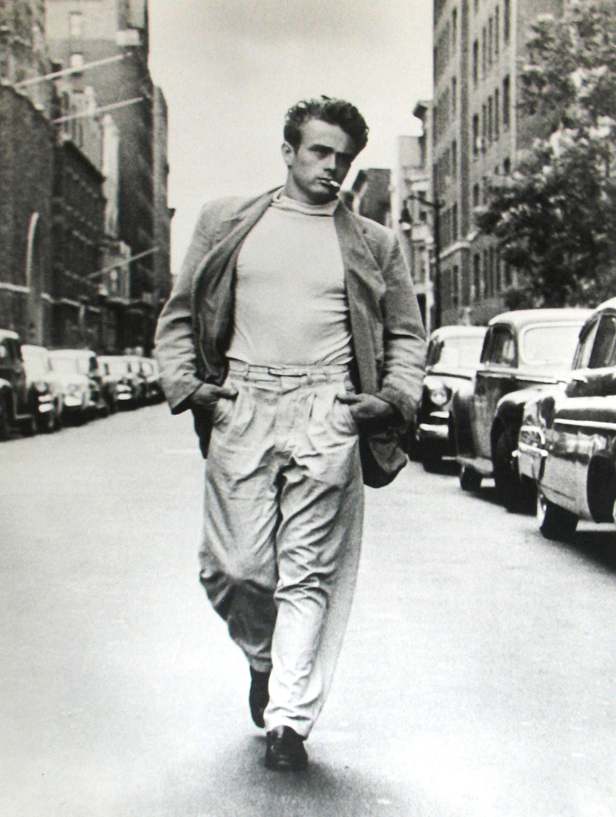 Black and white photograph of James Dean walking down a New York City street wearing a jacket, white shirt, and light pants, with a cigarette in his mouth