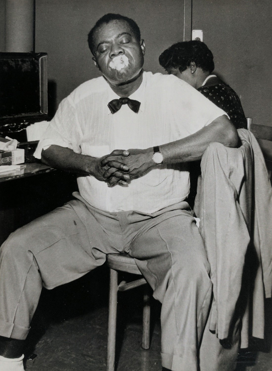 Black and white photograph of musician Louis Armstrong wearing a bowtie and sitting on a chair while making a funny face at the camera