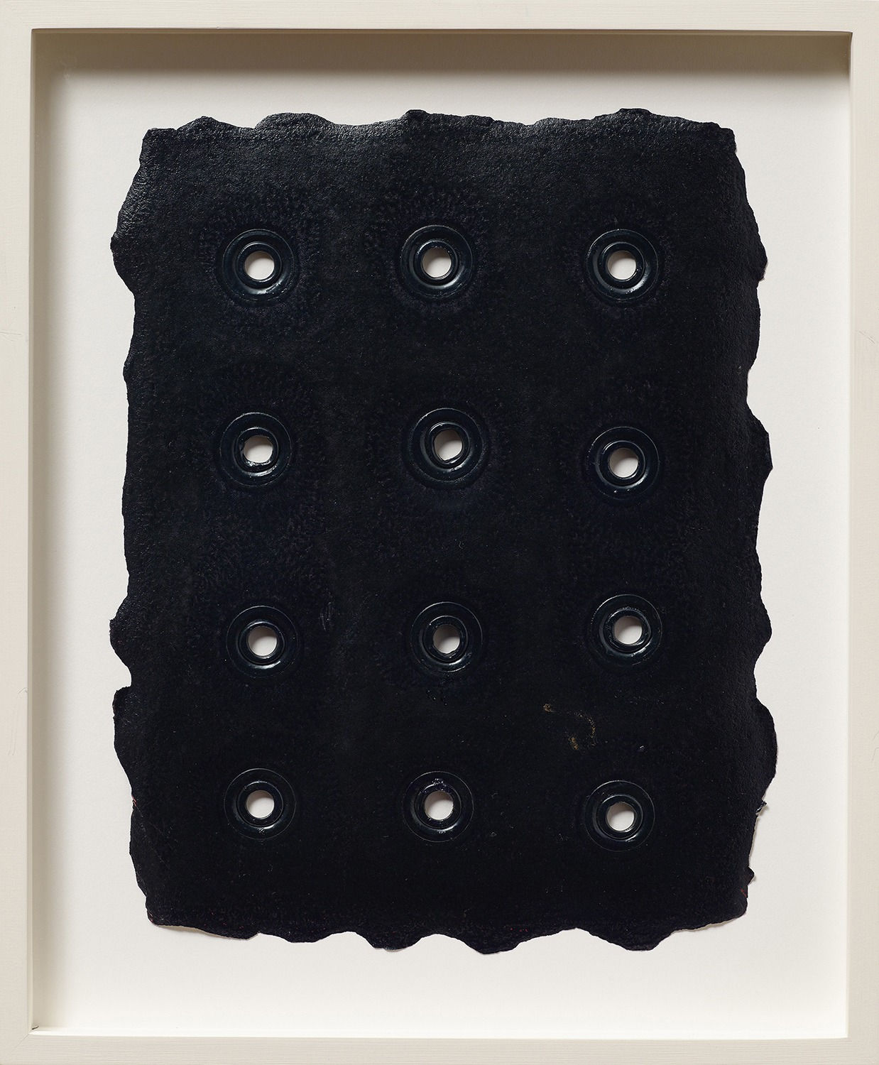 A framed piece of thick black paper with organic rough edges is penetrated by a neat array of twelve grommets