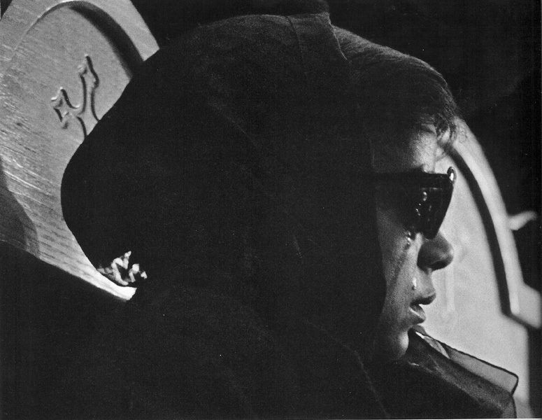Black and white photograph of Betty Shabazz, the widow of Malcolm X, in profile wearing a black headscarf and sunglasses with a streak of tears running down her cheek