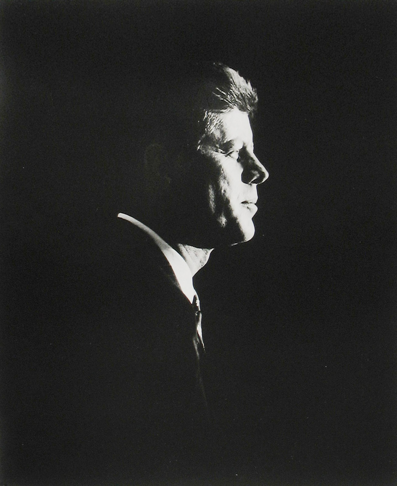 Black and white, high contrast photograph of John F Kennedy in profile against a pitch dark background