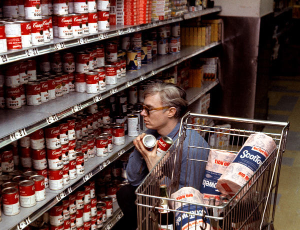 Photograph of Andy Warhol crouching in a grocery store aisle, selecting cans of soup behind a shopping cart