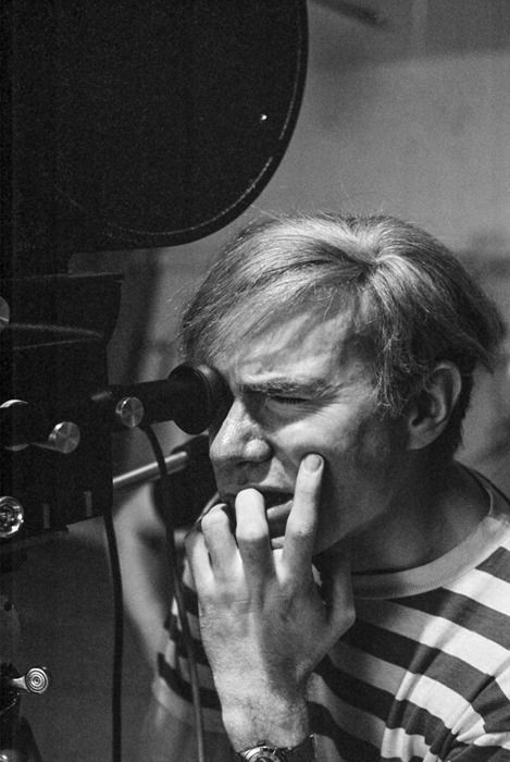 Black and white photograph of Andy Warhol looking into a film camera and biting his nails
