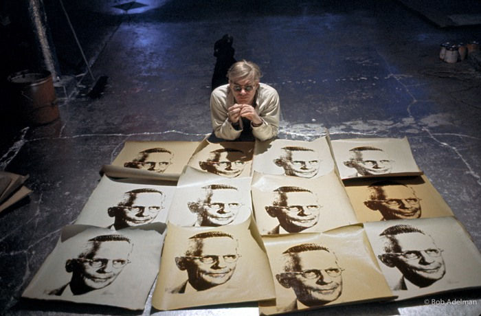 Photograph of Andy Warhol lying on his stomach looking at the viewer behind 12 screen prints laid on the floor